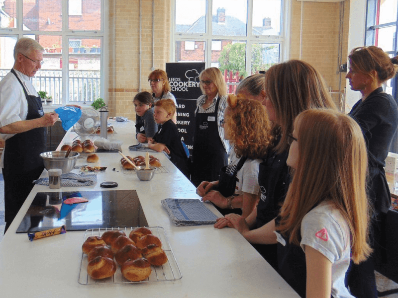 A man wearing chef's whites is teaching a group of adults and children how to make bread rolls